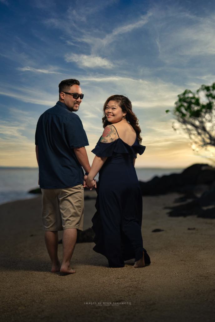 Oahu Engagement Photographer | Honolulu Beach Engagement Session A Honolulu engagement couple holds hands while walking on the beach at sunset with a view of the water and sky. Captured by a professional photographer on Oahu.