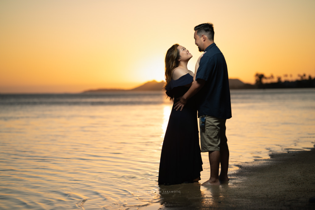Romantic Sunset Engagement Session at the Beach Couple, featuring an off-the-shoulder black dress and short-sleeved blue button-down, embraces during a sunset engagement shoot on an Oahu beach, with the mountain in view.