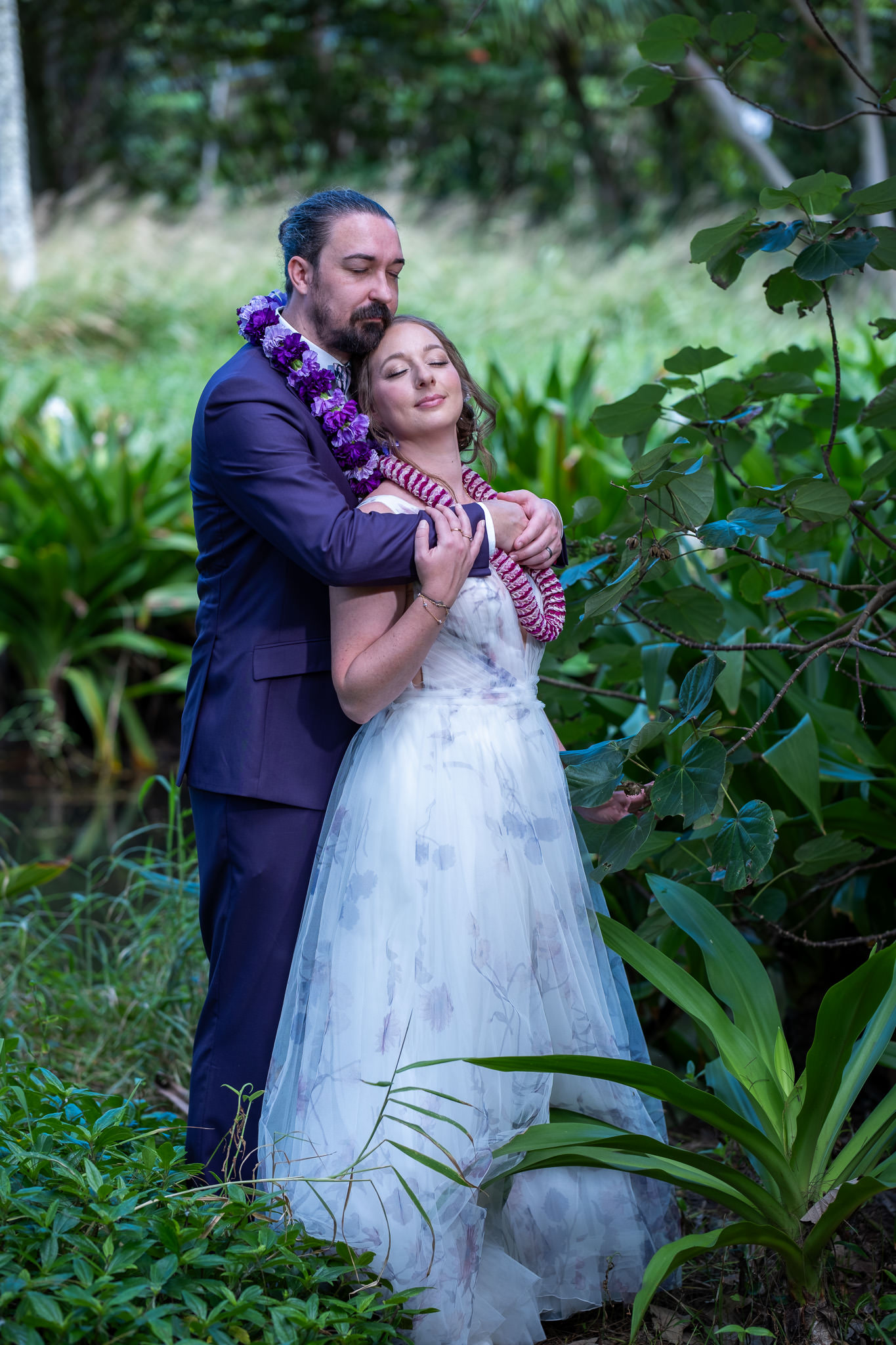 Honolulu Elopement Photography on Oahu An elopement photographer captures a couple in lei embracing in a lush Oahu forest. Professional Honolulu photography of a bride and groom in tropical greenery.