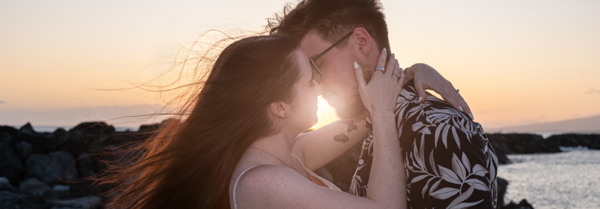Close-up of a couple kissing on a rocky beach at sunset during an Oahu creative photo session, with golden sunlight catching the woman's flowing hair.