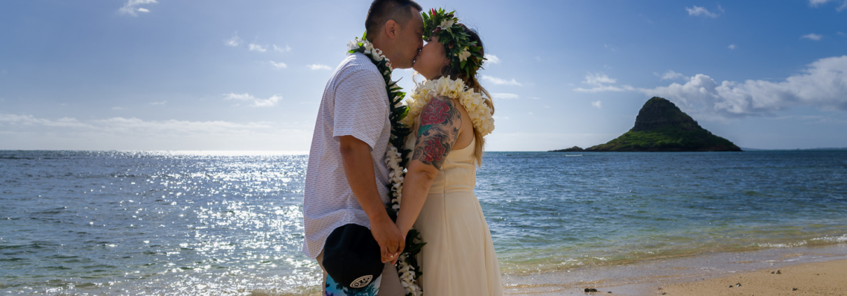 A newly married couple shares a romantic kiss at Kualoa Regional Park during their elopement wedding, with Mokoliʻi Island visible across the ocean.