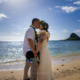 A newly married couple shares a romantic kiss at Kualoa Regional Park during their elopement wedding, with Mokoliʻi Island visible across the ocean.