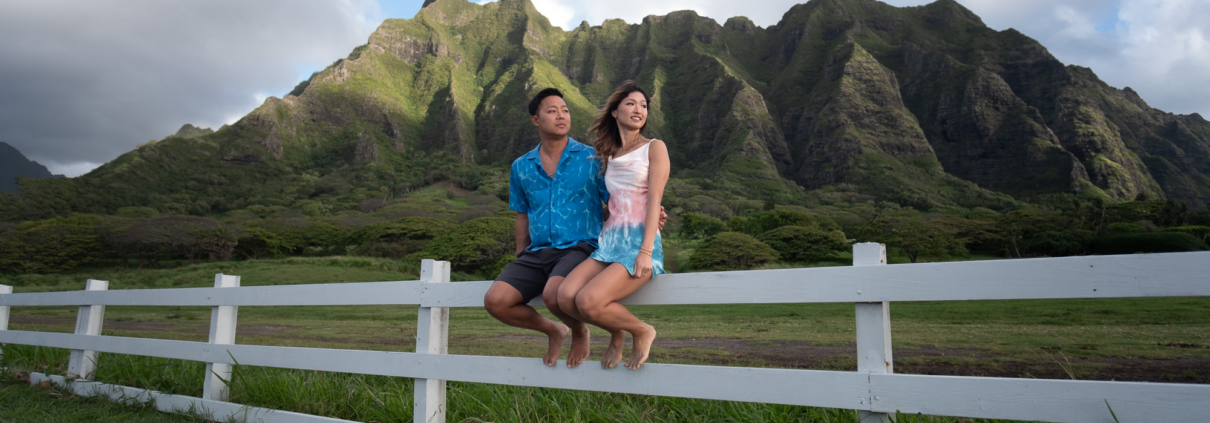 A scenic landscape photo from an Oahu ranch, capturing a couple casually sitting on a rustic white wooden fence bordering a wide, grassy field. They look over the meadow toward a dramatic mountain range.