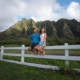 A scenic landscape photo from an Oahu ranch, capturing a couple casually sitting on a rustic white wooden fence bordering a wide, grassy field. They look over the meadow toward a dramatic mountain range.
