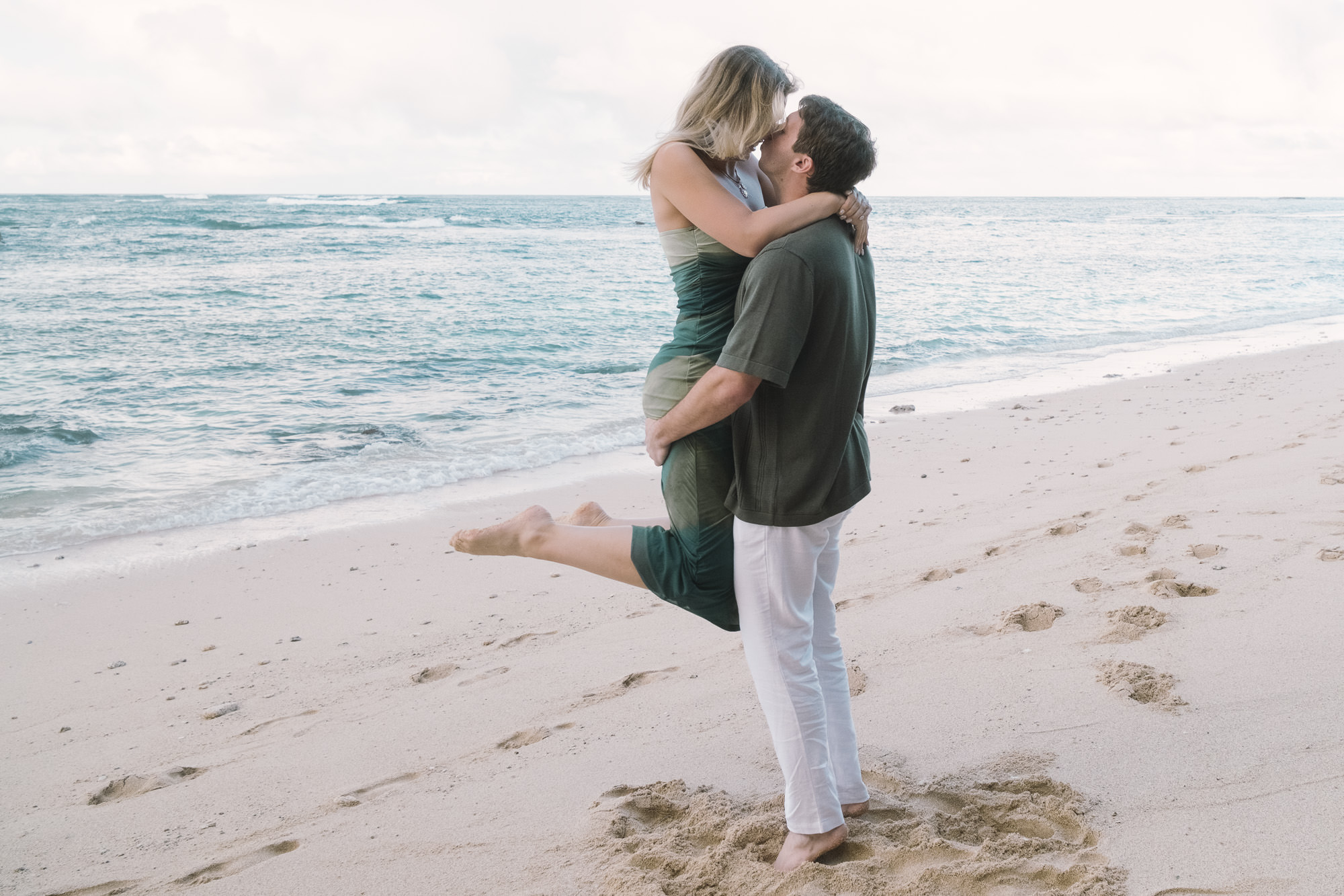 Man lifting his fiancée in his arms while kissing on the shoreline during an Oahu beach shoot.