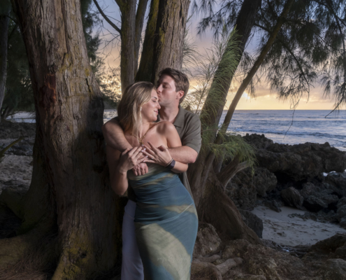 Man embracing his fiancée from behind and kissing her cheek under coastal trees on Oahu.