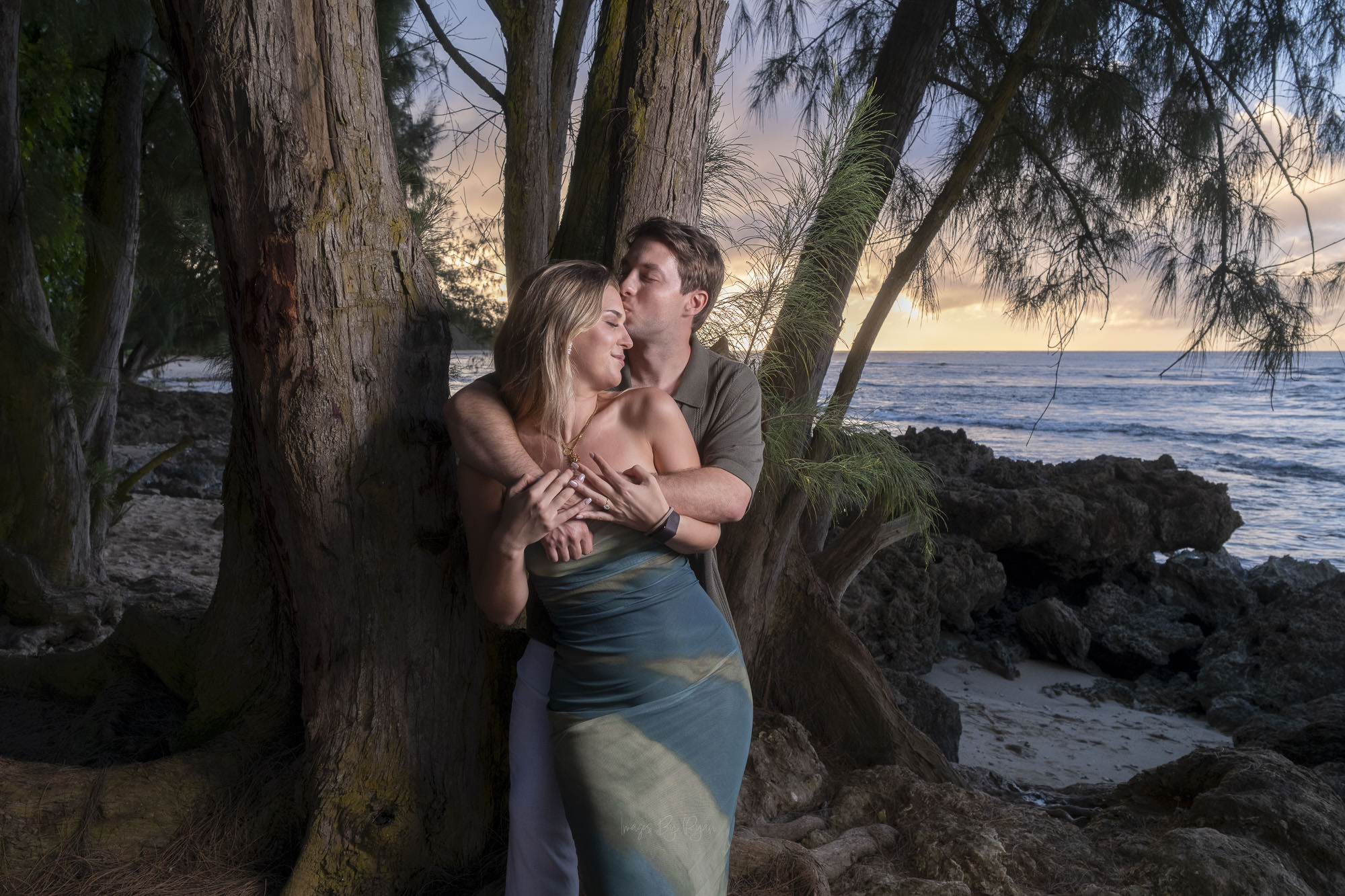Man embracing his fiancée from behind and kissing her cheek under coastal trees on Oahu.