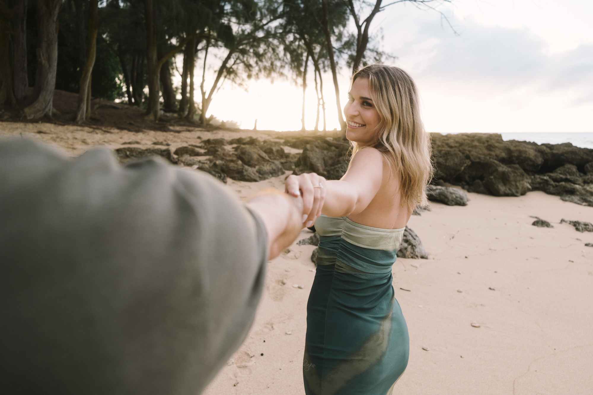 Woman in a green dress smiling while holding her partner's hand on an Oahu beach during a sunset proposal shoot.